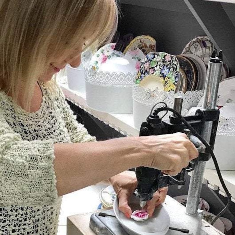 Pu&ograve; includere: Una persona usa una trapano a colonna per lavorare su un piccolo pezzo decorativo in ceramica. Il pezzo &egrave; bianco con un disegno floreale rosa. Scatole bianche con coperchio, finiture in pizzo e motivi floreali sono su uno scaffale.