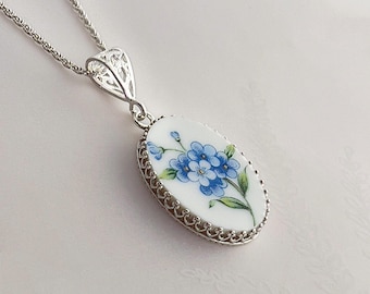 May include: Silver-toned necklace with an oval pendant featuring a floral design. The pendant has a white background with blue flowers and green leaves. The pendant is framed with a decorative silver border and hangs from a delicate chain.