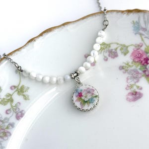 May include: A delicate necklace featuring a strand of white beads and a silver pendant. The pendant showcases a floral design with pink, blue, and green flowers. The necklace rests on a white plate with floral accents.