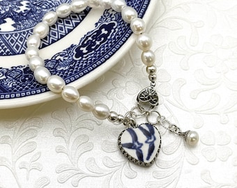 May include: A bracelet featuring a strand of white oval pearls and a silver-toned metal chain. The bracelet includes a heart-shaped charm with a blue and white floral pattern, a decorative silver heart connector, and a small dangling pearl accent.