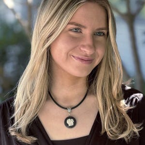 May include: A black circular pendant featuring a white relief design of a lion, suspended from a simple black cord necklace. The pendant is framed in a silver-toned metal setting with a decorative bail connecting it to the smooth black cord.