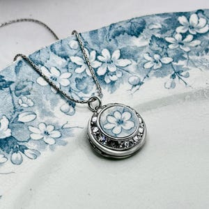 Puede incluir: Collar con colgante plateado con un dije redondo con un diseño floral. El dije presenta una flor blanca sobre un fondo azul, rodeada de cristales transparentes. La cadena del collar también es plateada.