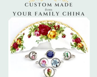 May include: A collection of silver rings with floral designs, displayed in front of a decorative china plate. The rings feature various colorful floral patterns. The text reads "CUSTOM MADE from YOUR FAMILY CHINA" and "Dinner Wear Jewelry".