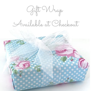 May include: A square gift box wrapped in light blue paper with white polka dots and pink rose floral patterns. A sheer white ribbon is tied in a bow on top. The text "Gift Wrap Available at Checkout" is written in cursive.