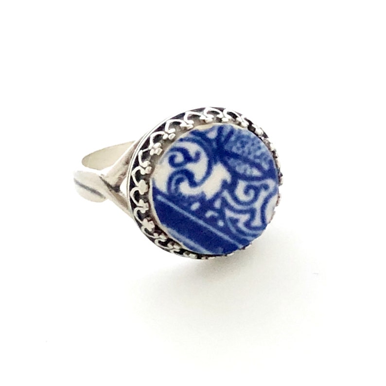 Pu&ograve; includere: Un anello d'argento con un elemento centrale rotondo in ceramica blu e bianca. La ceramica presenta un motivo decorativo. L'anello ha un design traforato attorno alla ceramica e una fascia semplice.