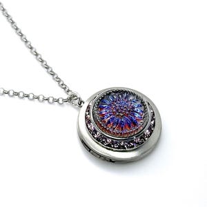 Puede incluir: Collar con colgante plateado de forma redonda. El colgante presenta un diseño floral de cristal en tonos azules, morados y rojos, rodeado de pequeños cristales brillantes. La cadena es fina.