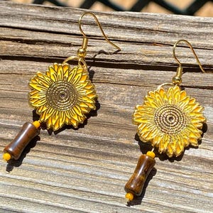 May include: A pair of gold-colored sunflower earrings with yellow enamel petals and a brown, cylindrical bead. The earrings have a gold hook and a small yellow bead at the base of the cylindrical bead.