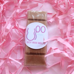 Peut inclure: Un paquet de fondants de cire de soja bruns avec une étiquette blanche qui indique "OO House SOY WAX MELTS". Les fondants sont enveloppés dans du plastique transparent et entourés de papier déchiqueté rose.