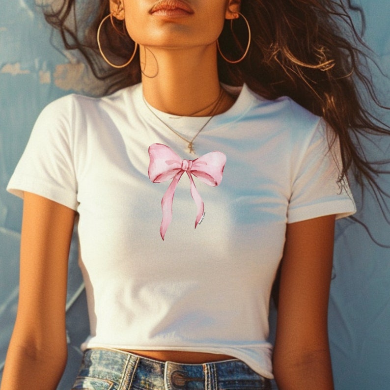 Pu&ograve; includere: T-shirt bianco con un disegno di fiocco rosa ad acquerello. Il fiocco ha lunghi nastri fluenti ed &egrave; centrato sul davanti. La maglietta ha maniche corte e girocollo.
