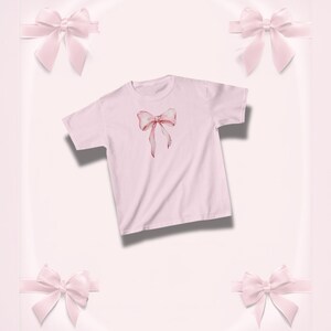 Pu&ograve; includere: Una maglietta rosa chiaro con un disegno di fiocco ad acquerello nei toni del rosa e del rosso. Il fiocco &egrave; centrato sulla maglietta. Lo sfondo &egrave; rosa chiaro con quattro fiocchi rosa decorativi.