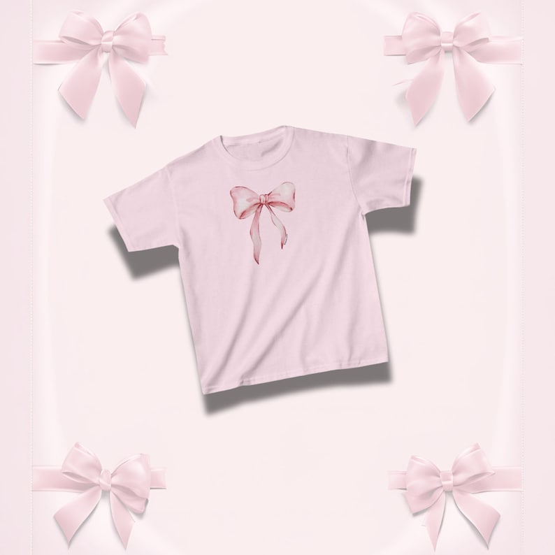 Pu&ograve; includere: Una maglietta rosa chiaro con un disegno di fiocco ad acquerello nei toni del rosa e del rosso. Il fiocco &egrave; centrato sulla maglietta. Lo sfondo &egrave; rosa chiaro con quattro fiocchi rosa decorativi.