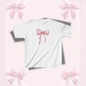 Pu&ograve; includere: T-shirt bianco con un design a fiocco rosa in stile acquerello. Il fiocco ha nastri fluenti ed &egrave; centrato sulla maglietta. Lo sfondo &egrave; rosa tenue con ulteriori accenti di fiocco rosa.