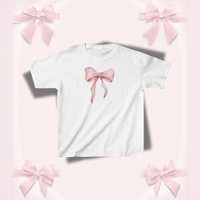 Pu&ograve; includere: T-shirt bianco con un design a fiocco rosa in stile acquerello. Il fiocco ha nastri fluenti ed &egrave; centrato sulla maglietta. Lo sfondo &egrave; rosa tenue con ulteriori accenti di fiocco rosa.