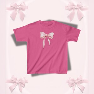 Pu&ograve; includere: Una maglietta rosa acceso con un disegno di fiocco ad acquerello. Il fiocco &egrave; rosa chiaro con sfumature delicate e nastri lunghi e fluenti. La maglietta &egrave; di colore pieno, con girocollo classico e maniche corte. Lo sfondo presenta fiocchi rosa decorativi.