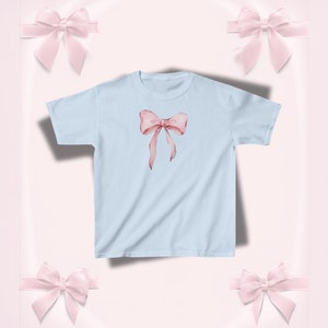 Pu&ograve; includere: T-shirt azzurra con un motivo a fiocco rosa acquerellato. Il fiocco, con lunghi nastri fluttuanti, &egrave; centrato sul davanti. La maglietta &egrave; in stile girocollo classico.