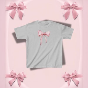 Pu&ograve; includere: Una maglietta grigio chiaro con un design a fiocco rosa in stile acquerello. Il fiocco ha lunghi nastri fluenti. Lo sfondo &egrave; rosa tenue con fiocchi rosa decorativi.