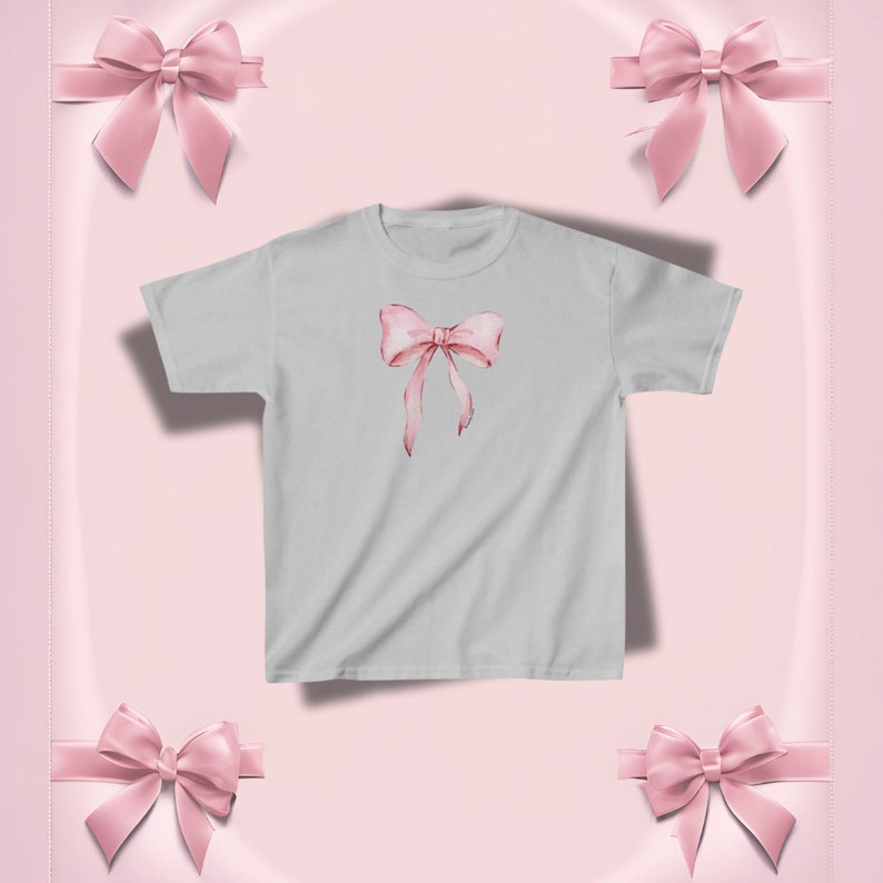 Pu&ograve; includere: Una maglietta grigio chiaro con un design a fiocco rosa in stile acquerello. Il fiocco ha lunghi nastri fluenti. Lo sfondo &egrave; rosa tenue con fiocchi rosa decorativi.
