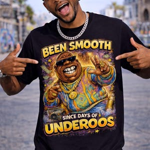 May include: Black t-shirt with a colorful graphic of a cartoon character wearing sunglasses and gold jewelry. The text "BEEN SMOOTH SINCE DAYS OF UNDEROOS" is printed in yellow. The person in the image is pointing at the shirt.