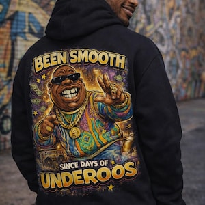 May include: Black hoodie featuring a colorful cartoon illustration of a man wearing sunglasses and gold jewelry. The text "BEEN SMOOTH SINCE DAYS OF UNDEROOS" is printed above and below the illustration. The hoodie is worn by a person.