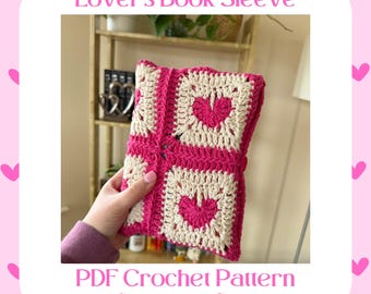 Puede incluir: Una funda de libro de crochet rosa y blanca con un diseño de corazón. La funda está siendo sostenida por una mano. El texto "Lover's Book Sleeve" y "PDF Crochet Pattern BY STITCHED & CRAFTED" es visible.
