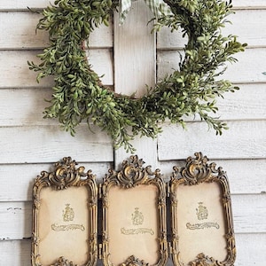 May include: A triptych of three ornate, gold-toned metal picture frames with intricate scrollwork detailing, each containing a beige insert with a crest logo. Above the frames hangs a circular wreath of green leaves tied with a green and white gingham ribbon.