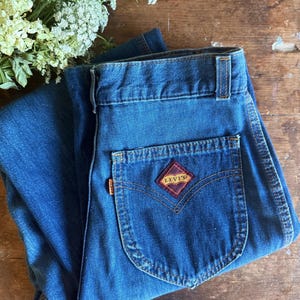 May include: Medium blue denim jeans featuring a high-waisted design with belt loops and a single back pocket. The pocket displays a diamond-shaped patch with a red plaid pattern and the Levi's brand name in gold text, plus an orange side tab.