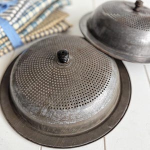 May include: Two vintage metal colanders with a perforated design and a small black knob on top. The colanders are gray and have a rustic, aged appearance. A stack of blue and white checkered fabric napkins is in the background.