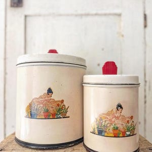 May include: Two vintage cream-colored metal canisters with black trim and red knobs. Each canister features a colorful illustration of a woman tending to potted plants. The canisters are displayed on a wooden surface.