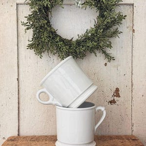 May include: Two white ceramic mugs with a smooth, glossy finish and a distinct base rim. One mug is positioned upright on a wooden surface, while the second mug is stacked at an angle on top of the first, featuring a curved handle on each side.