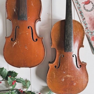 May include: Two antique violins with a warm, aged appearance hang against a white background. The violins have a reddish-brown finish with visible wear and tear. A Christmas-themed book and holly sprigs with red berries add festive accents.