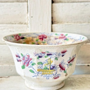 May include: A ceramic bowl with a cream-colored base and a visible crackled glaze finish. The exterior features a colorful floral pattern with pink, purple, and green flowers surrounding a central yellow basket motif, with similar floral designs inside.