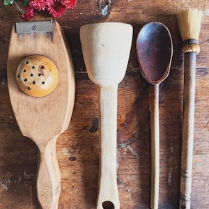 May include: A collection of vintage wooden kitchen utensils, including a butter paddle with a metal blade, a mortar, a spoon, and a brush. The items are arranged on a rustic wooden surface, with red flowers in the background.