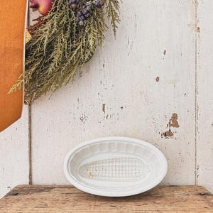 May include: An oval-shaped white ceramic mold featuring a molded corn cob design at the center. The dish has a fluted rim and a smooth, glossy finish, presenting a shallow basin structure suitable for shaping food items or as a decorative piece.