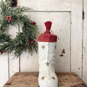 May include: A decorative Christmas stocking in a rustic style. The stocking is white with gold stars and a red top. It is sitting on a wooden surface with a wreath in the background.