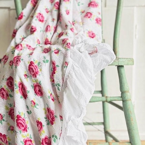 May include: A white floral blanket with a pattern of pink roses and green leaves is draped over a distressed, light green wooden chair. The blanket has a ruffled white trim.