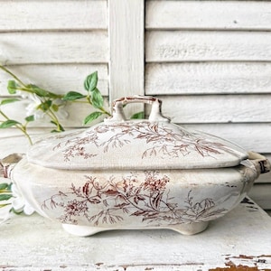 May include: Cream-colored antique serving dish with a lid, featuring a floral pattern in shades of brown. The dish has a handle on the lid and small feet. The background is a white, weathered wooden surface.