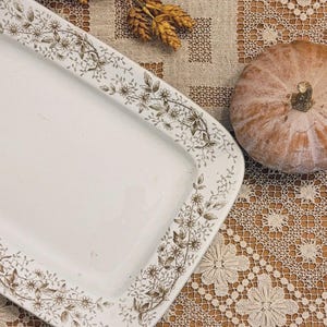 May include: A rectangular white ceramic serving platter featuring a brown floral vine pattern along the rim. The platter is positioned next to a small orange pumpkin and dried wheat stalks on a beige lace tablecloth.