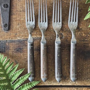 May include: Four antique forks with wooden handles and metal tines are arranged on a wooden surface. The handles have a dark brown finish with a metal accent at the base. The forks are vintage and show signs of wear.