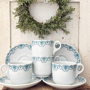 May include: A set of three white ceramic teacups and three matching saucers, each decorated with a teal blue floral and scroll pattern along the rim. The cups feature a rounded handle and a smooth finish, arranged in a stacked and side-by-side display.