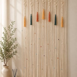 May include: Cream macrame wall hanging with a wooden rod. It features diamond patterns, tassels in yellow, teal, and orange, and small, colourful beads. This decorative piece adds a bohemian touch to any room.