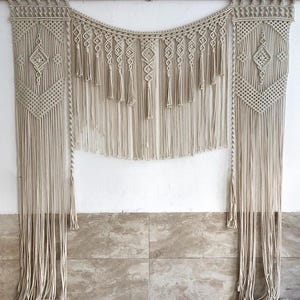 May include: A macrame wall hanging with a geometric design in a cream color. The hanging is made of cotton cord and has a fringe at the bottom.