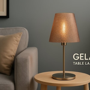 Könnte beinhalten: Eine Tischlampe mit einem strukturierten, pfirsichfarbenen Lampenschirm und einem goldfarbenen Sockel und Stiel. Die Lampe steht auf einem runden, hellen Holztisch. Der Text "GELA TABLE LAMP" ist sichtbar.