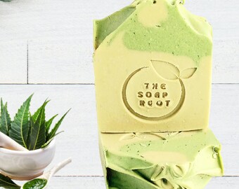 May include: Two bars of handmade soap with a light green and yellow swirl pattern. The soap has the text "THE SOAP ROOT" stamped on the front. Fresh green leaves and a mortar and pestle are in the background.