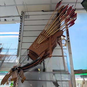 Puede incluir: Un carcaj de tiro con arco de cuero marrón lleno de flechas, algunas con plumas rojas. El carcaj está unido a un arco. El carcaj tiene costuras decorativas y remaches de latón. El arco es de madera y tiene una empuñadura negra.
