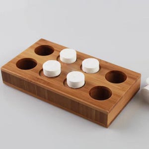 Puede incluir: Una bandeja de madera con ocho compartimentos redondos, cinco de los cuales están llenos de almohadillas redondas de algodón blanco para desmaquillar. La bandeja está hecha de bambú marrón claro.