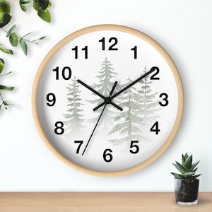 May include: A round wall clock with a light wood frame. The clock face is white with black numbers and hands. The center features a watercolor painting of evergreen trees in shades of green and gray. A small succulent plant in a black pot is visible.