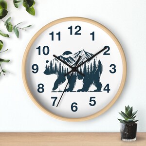 May include: A white clock with black numbers and hands, featuring a silhouette of a bear in a forest setting. The bear is outlined in blue and the forest is filled with trees. The clock has a wooden frame.