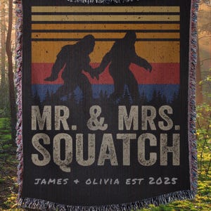 May include: Black woven throw blanket with a Bigfoot couple silhouette design. The blanket features a retro sunset color scheme and the text "MR. & MRS. SQUATCH" with the names "JAMES + OLIVIA EST 2025". The blanket has a fringed edge.