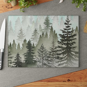 May include: Rectangular cutting board with a watercolor forest scene. The design features layers of green and gray pine trees against a light blue sky. The board is made of glass and is suitable for kitchen use.