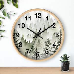 May include: Round wall clock with a light wood frame. The clock face features a watercolor painting of a misty forest in shades of green and gray. Black hands and numerals complete the design, offering a nature-inspired aesthetic.